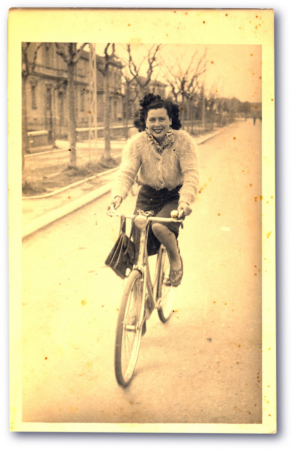 historische etwas vergilbte Postkarte: Auf einer leeren Straße mit großen Häusern und blätterlosen Bäumen im Hintergrund fährt eine Frau mit dunklen Locken, knielangem Rock und Strickjacke auf einem Fahrrad, sie lacht 